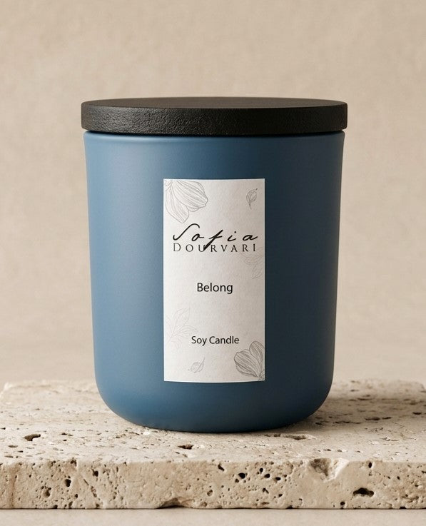 Sofia Dourvari Belong soy scented candle in a matte dusty blue vessel with black lid, displayed on a travertine stone surface