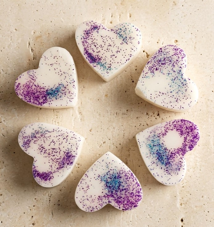 Sofia Dourvari Black Fig & Vetiver heart-shaped coconut wax melts with purple and blue shimmer swirl, arranged in a circle on a travertine stone