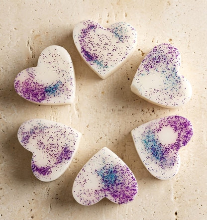 Sofia Dourvari Black Fig & Vetiver heart-shaped coconut wax melts with purple and blue shimmer swirl, arranged in a circle on a travertine stone