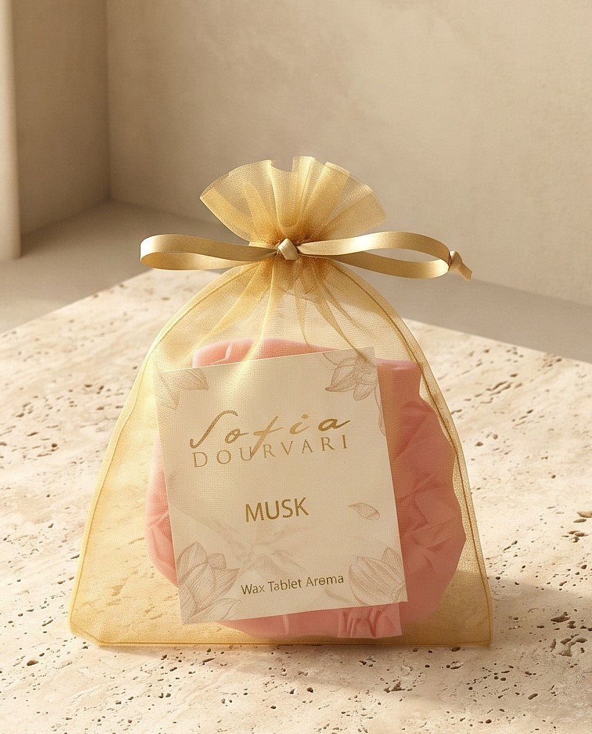 Sofia Dourvari Musk Wax Tablet Aroma in a tied gold organza bag with gold-script branded label, pink wax tablets visible inside, on a travertine stone surface