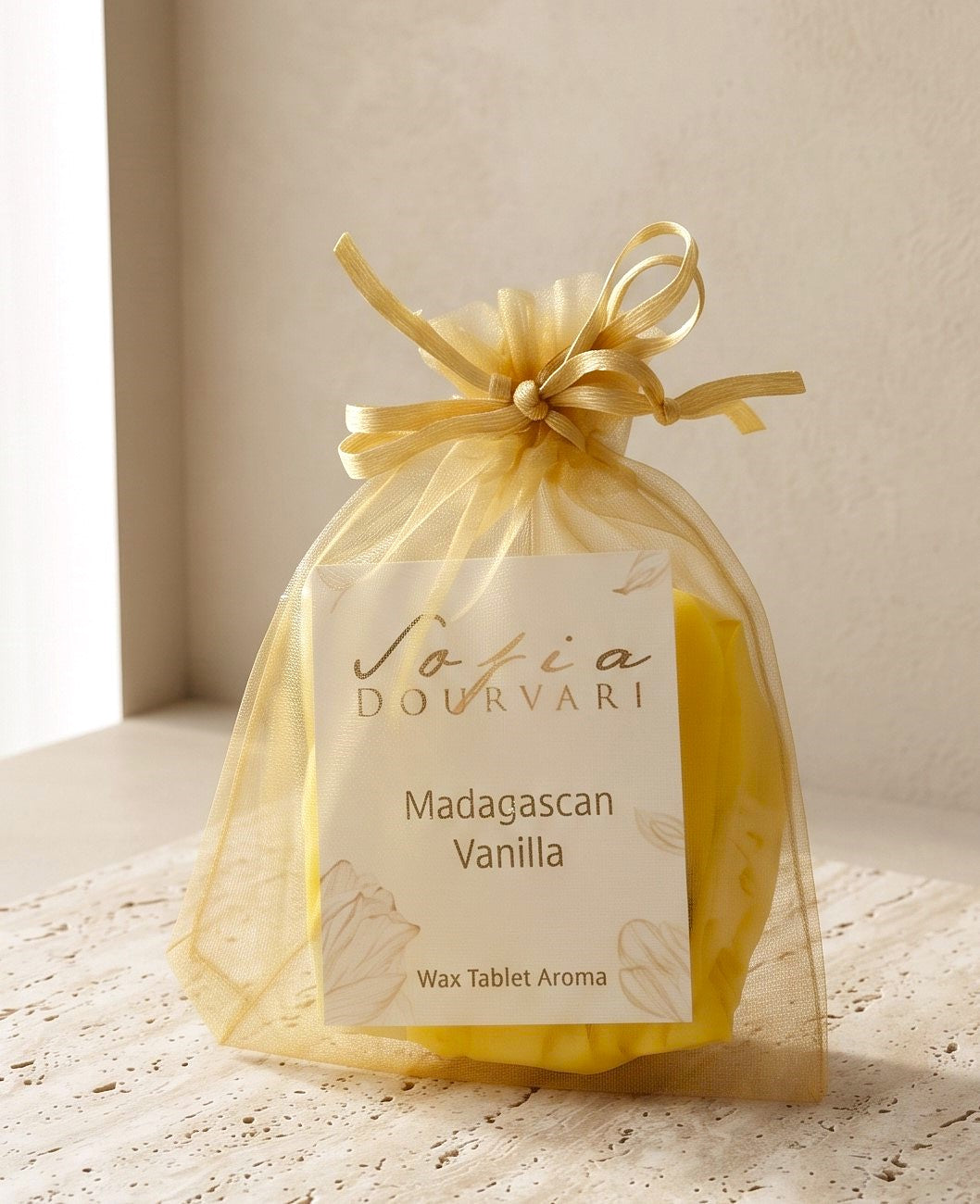 Sofia Dourvari Madagascan Vanilla Wax Tablet Aroma in a tied gold organza bag with branded label, yellow wax tablets visible inside, in soft window light on a travertine surface