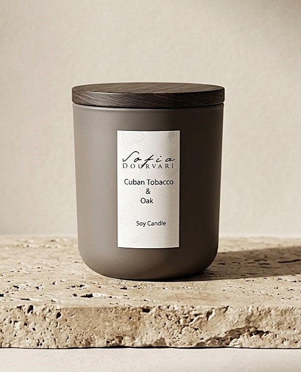 Sofia Dourvari Cuban Tobacco & Oak soy scented candle in a matte taupe vessel with dark wooden lid, on a travertine stone surface