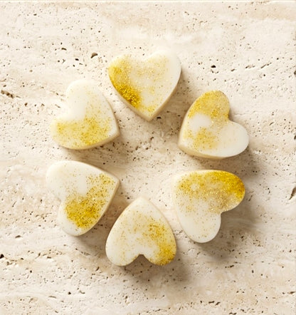 Sofia Dourvari Madagascan Vanilla heart-shaped coconut wax melts with gold shimmer detailing, arranged in a circle on a travertine stone surface